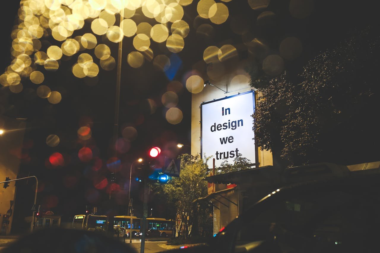 A city street at night featuring an illuminated billboard with the phrase 'In design we trust.'